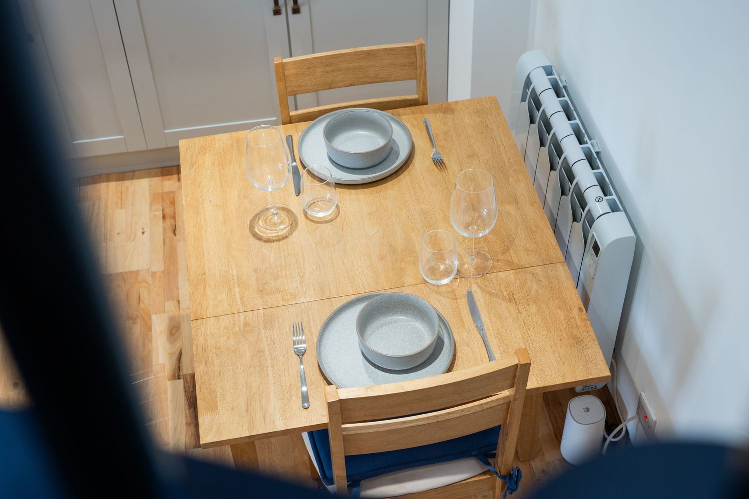 Wooden dining table set for two with plates and wine glasses near a white wall-mounted radiator.