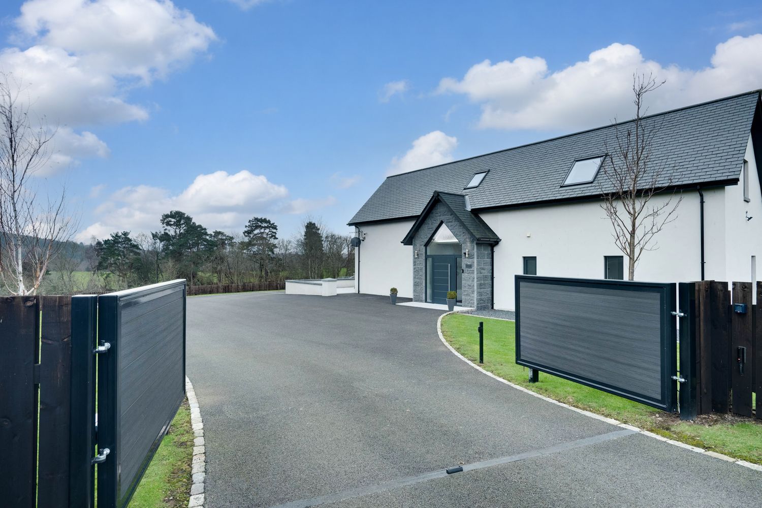 Modern white house with gray roof and sliding entrance gates on curved driveway under blue sky with puffy clouds.