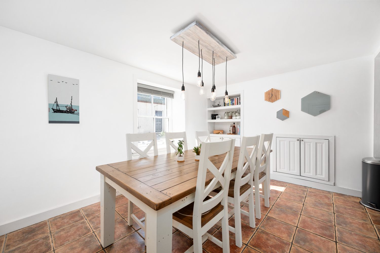 Modern white dining room with rustic wooden table, cross-back chairs, and pendant lighting over terra cotta flooring.