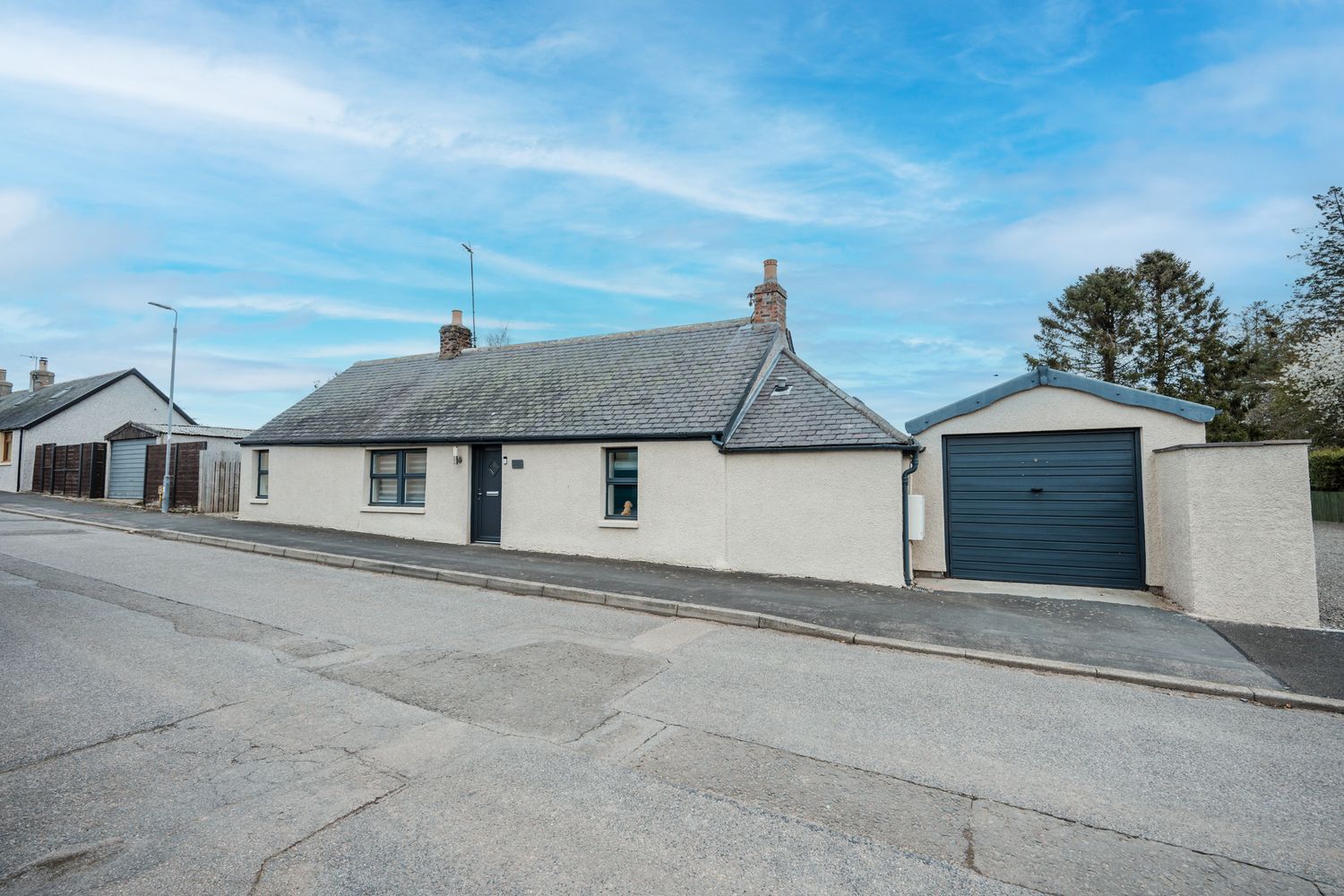 White rural cottage style house with blue garage door on quiet residential street under bright blue sky.