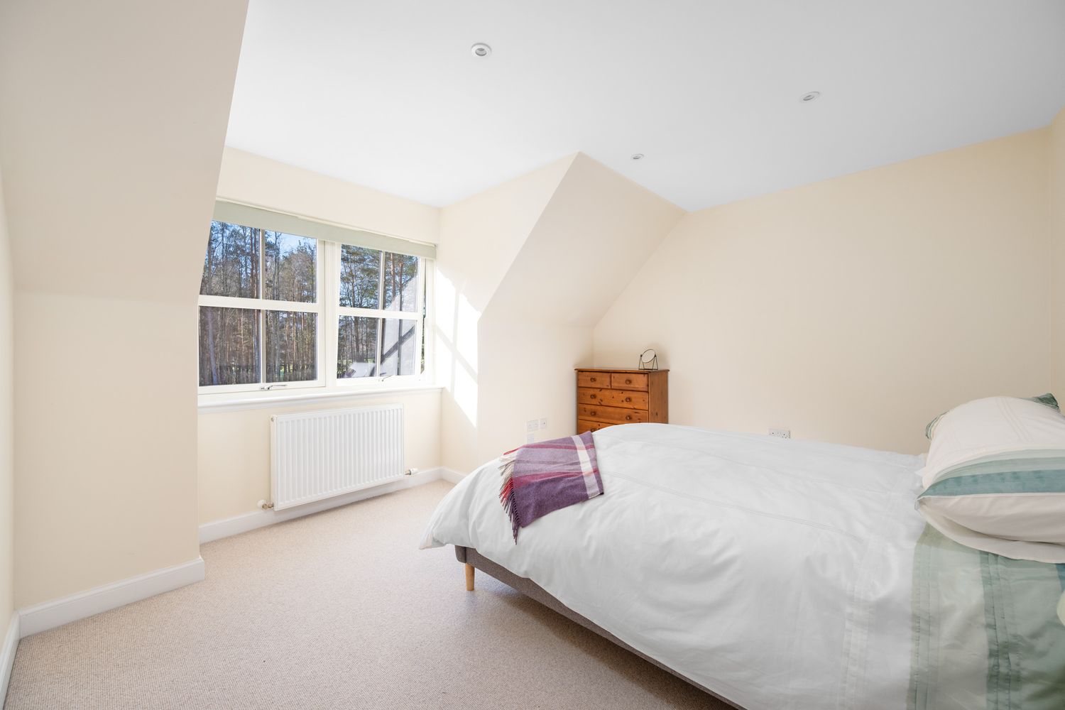 Bright and airy bedroom with sloped ceiling, white bedding, beige walls, and a window letting in natural light.