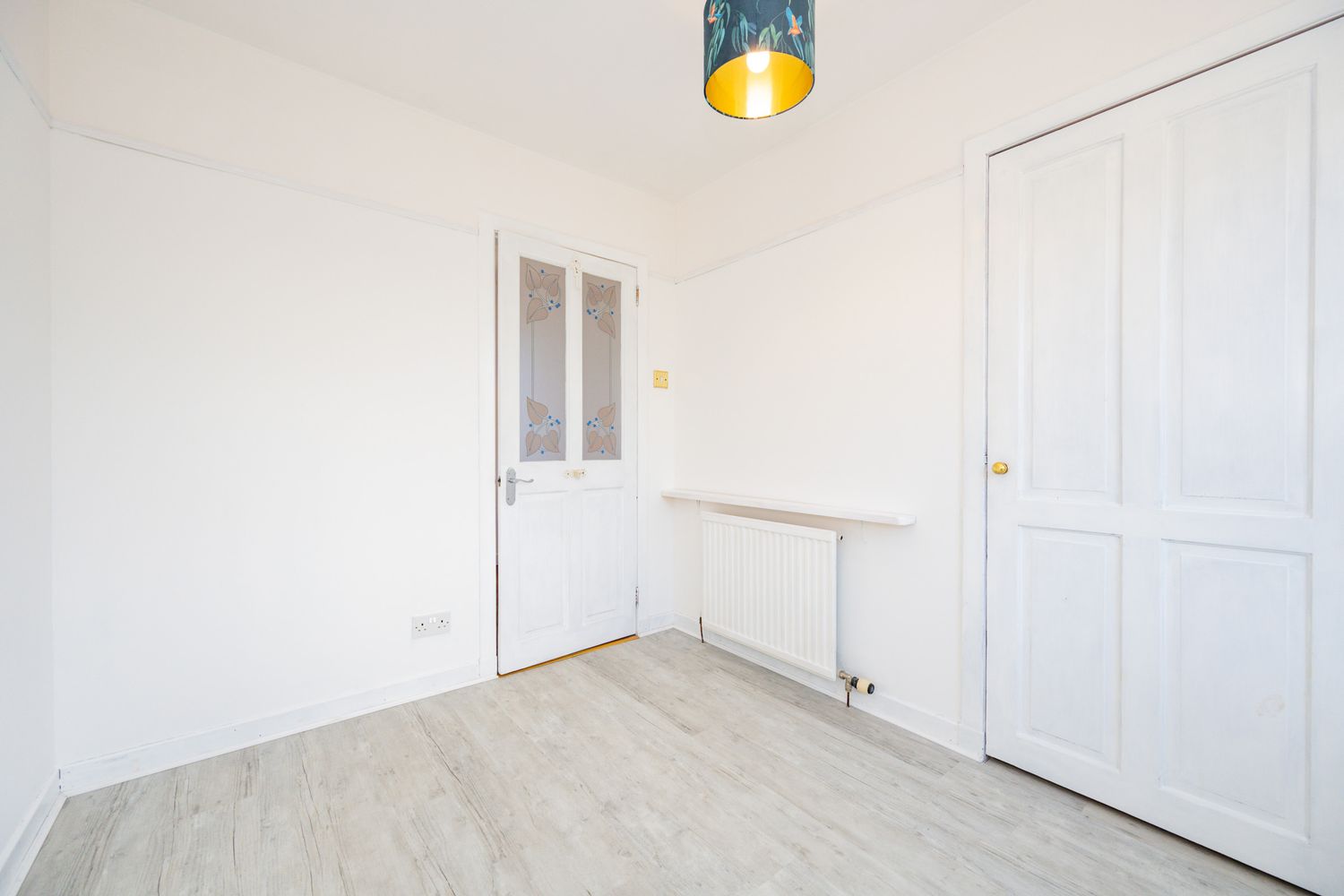 Empty white room with light wood flooring, white walls, radiator, and two white doors under a pendant light fixture.