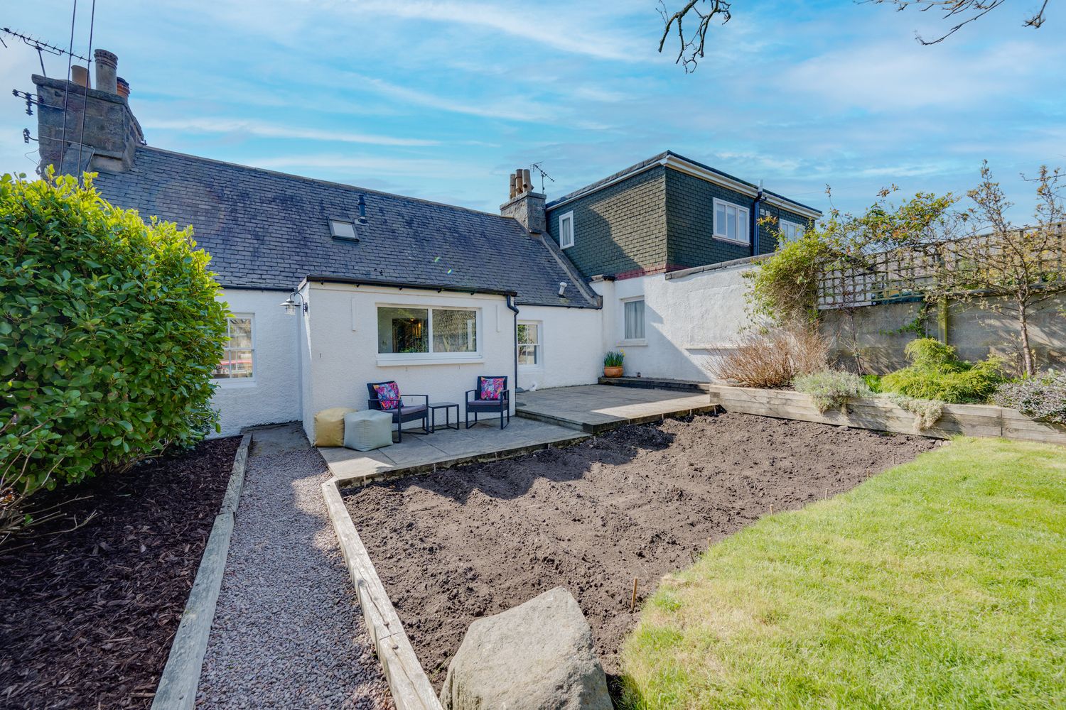 White cottage house with deck and landscaped backyard featuring freshly tilled soil and manicured lawn on sunny day.