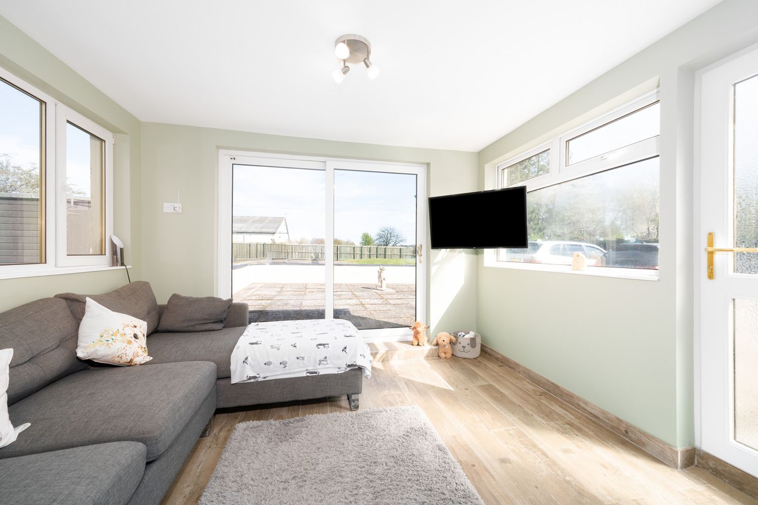 Modern living room with grey sectional sofa, hardwood floors, large windows, and a wall-mounted TV offering bright views.