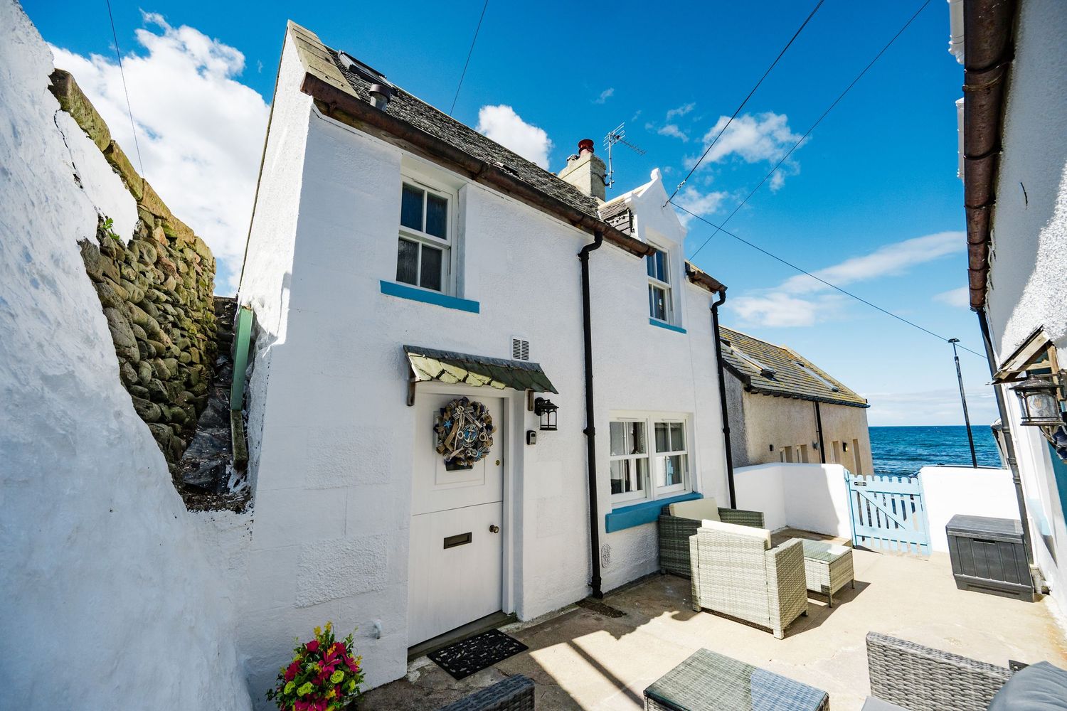Charming white coastal cottage with blue trim and a wreath on the door overlooking the sea on a sunny day.