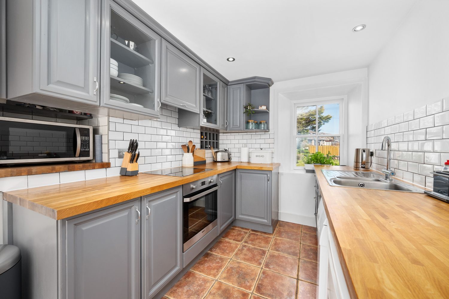 Modern galley kitchen with gray cabinets, butcher block countertops, subway tile backsplash and terracotta flooring.