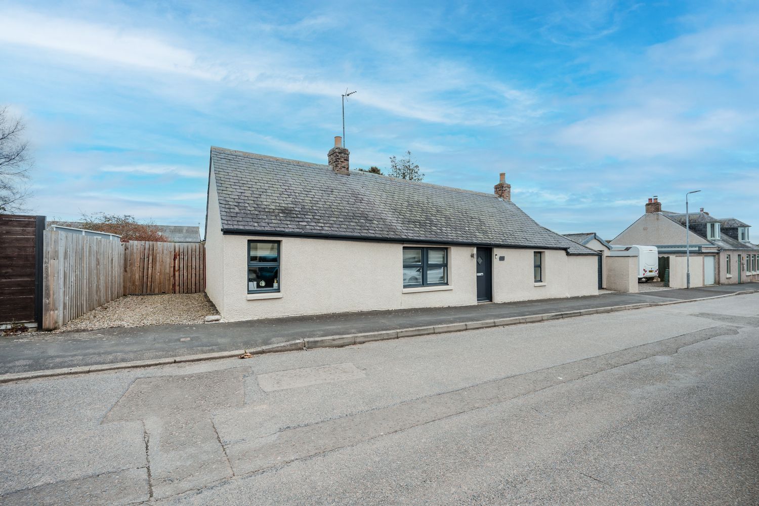 Traditional white Scottish cottage with gray roof along a quiet residential street under bright blue sky in rural village.