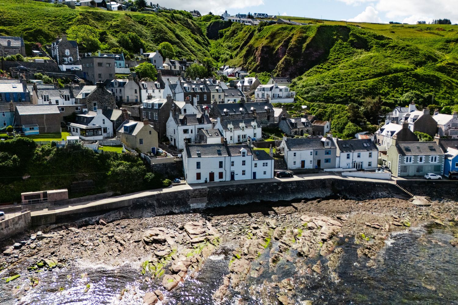 Scenic coastal village with white houses nestled on green hillside overlooking rocky shoreline in Scotland.