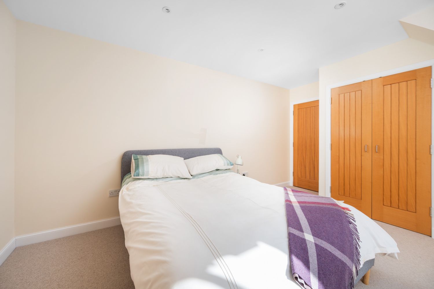 A small bedroom featuring a white bed with purple throw blanket and wooden closet doors in neutral tones.