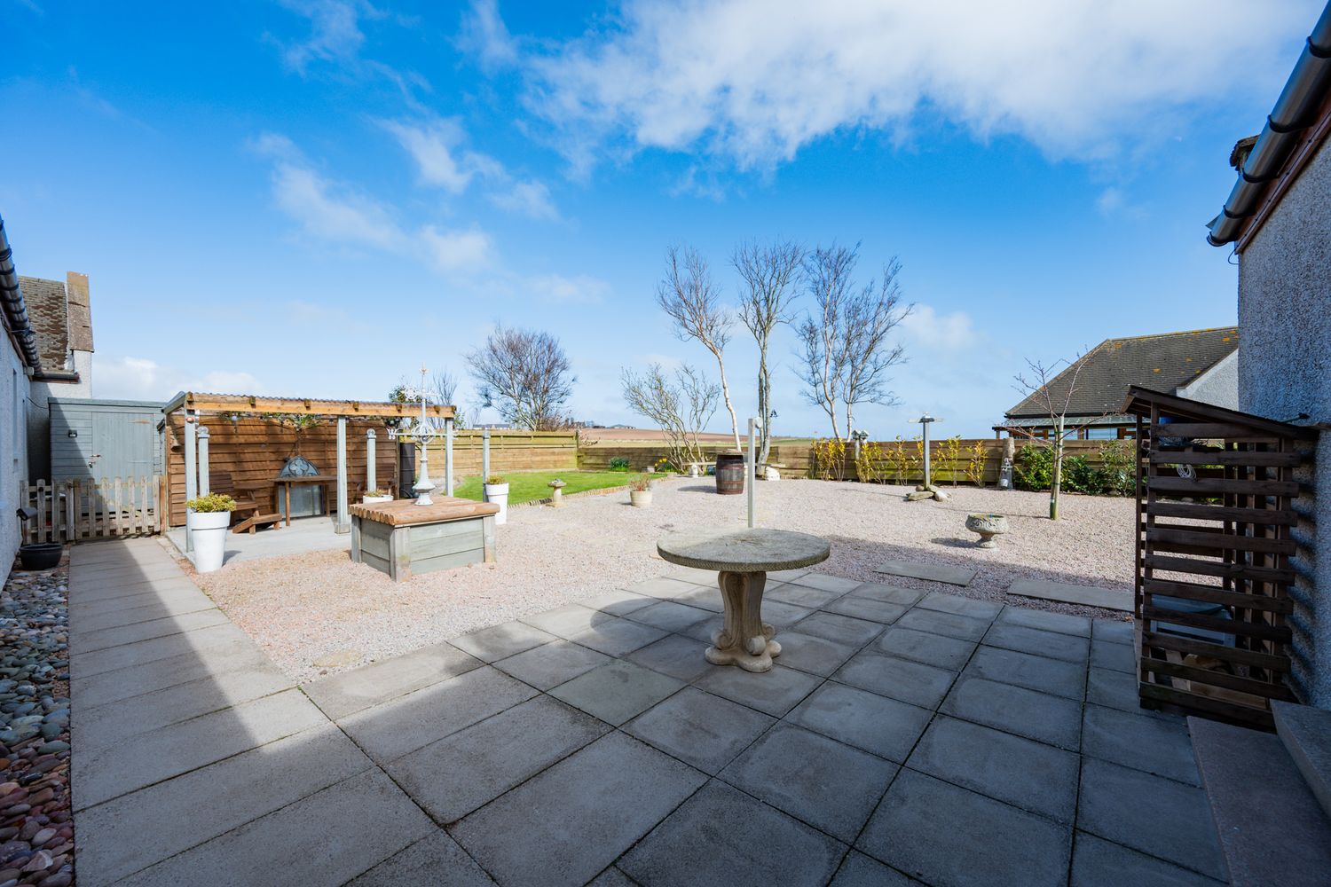 Spacious stone paved patio with wooden pergola and birdbath feature overlooking scenic countryside views on sunny day.