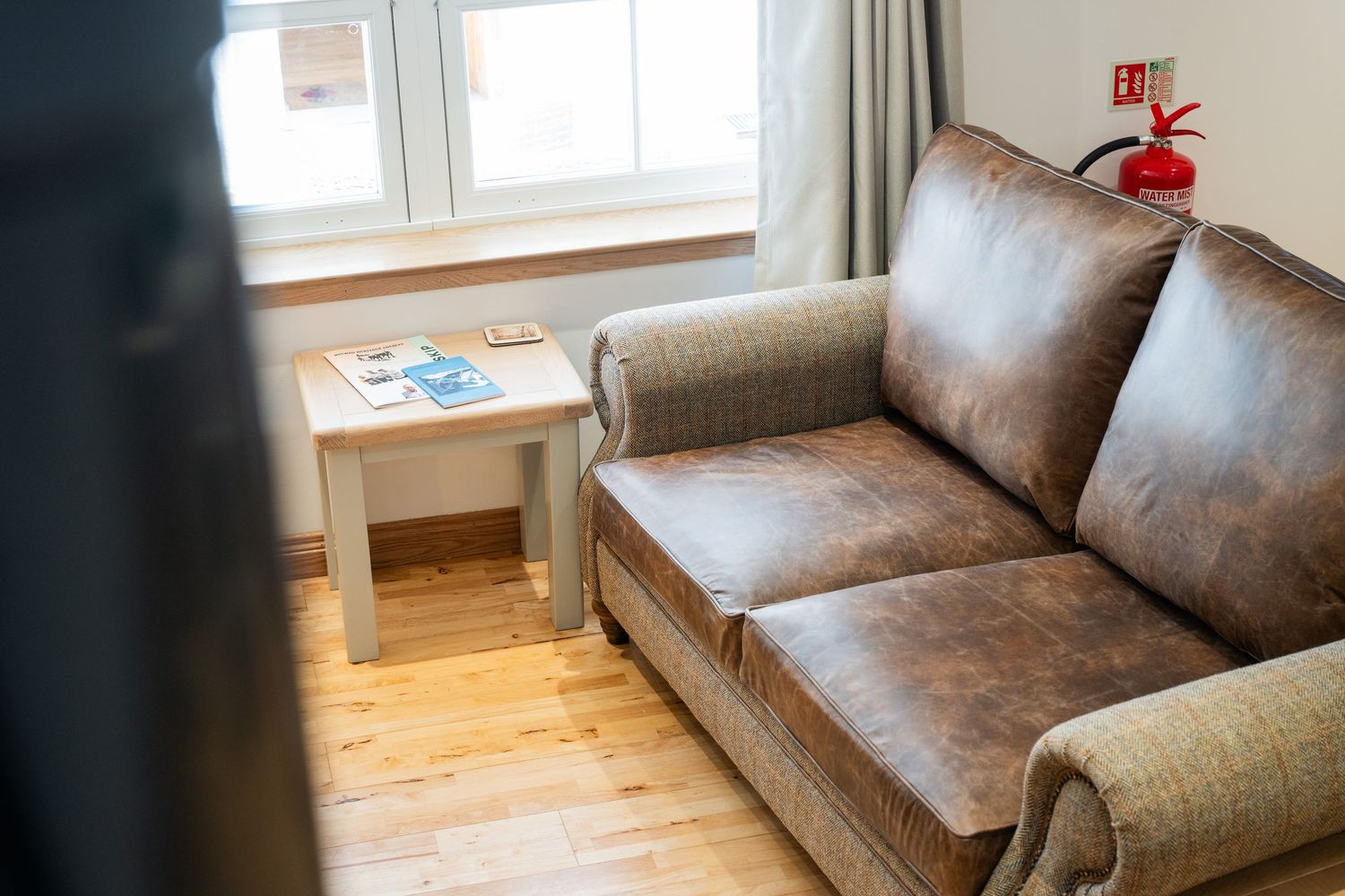 Brown leather couch with throw pillow next to window and small side table in cozy living room interior.