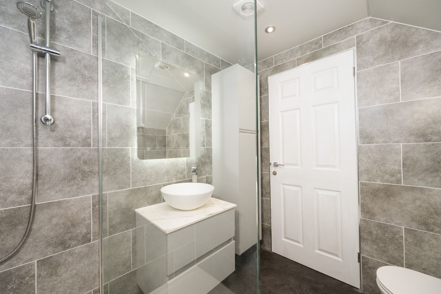 Modern bathroom interior with gray marble tiles, floating white vanity, illuminated mirror and glass shower enclosure.