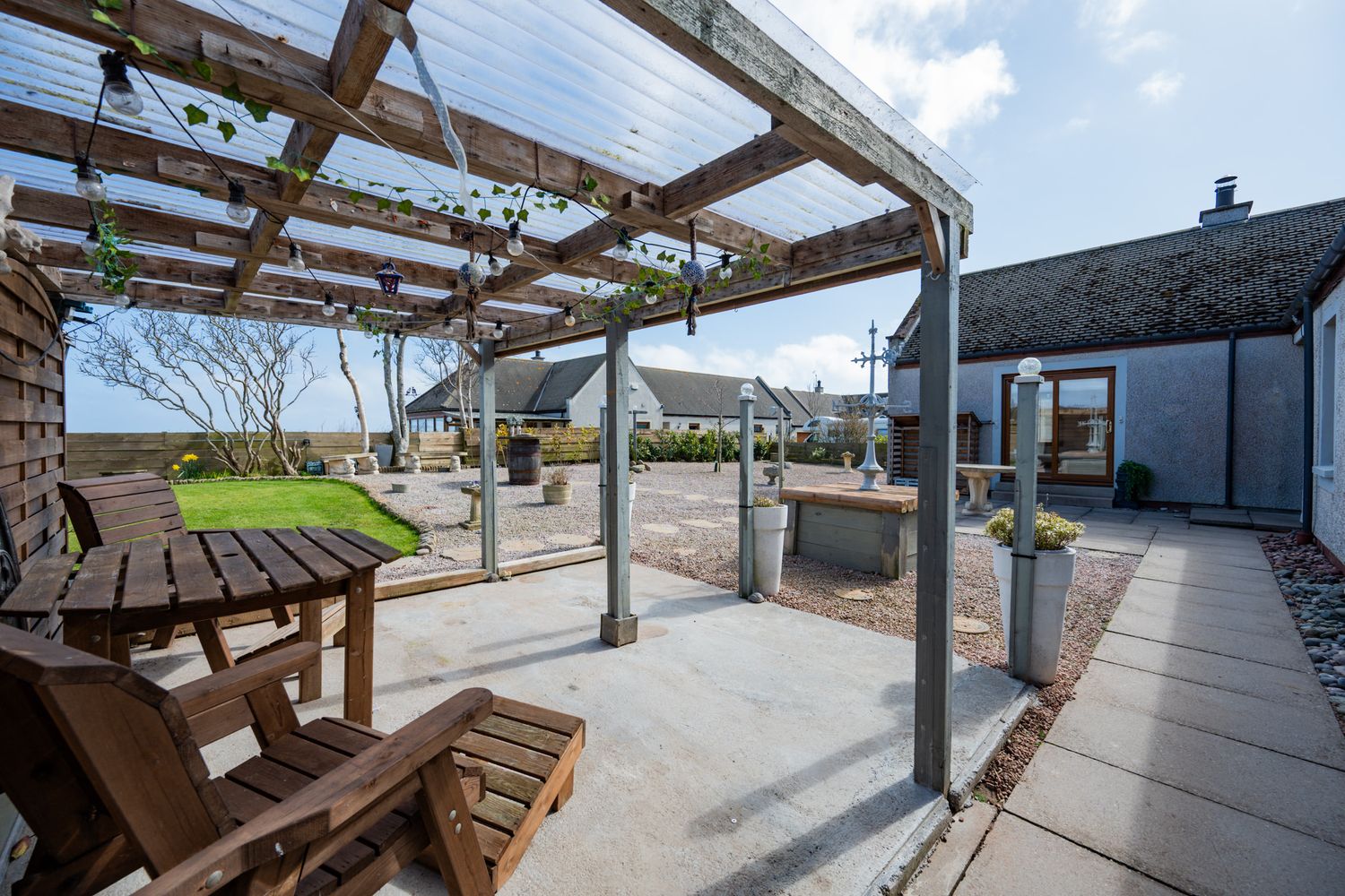 Covered outdoor patio with wooden pergola, picnic table and built-in grill overlooking a backyard on a sunny day.