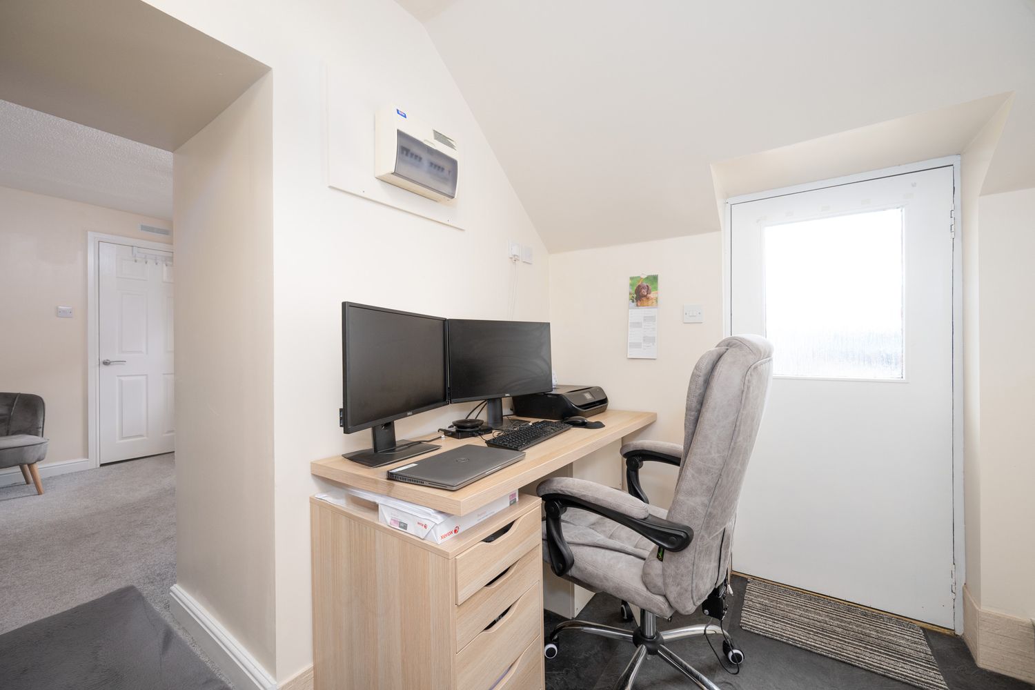 A home office setup with dual monitors on a wooden desk, grey office chair, and bright white walls with a window.