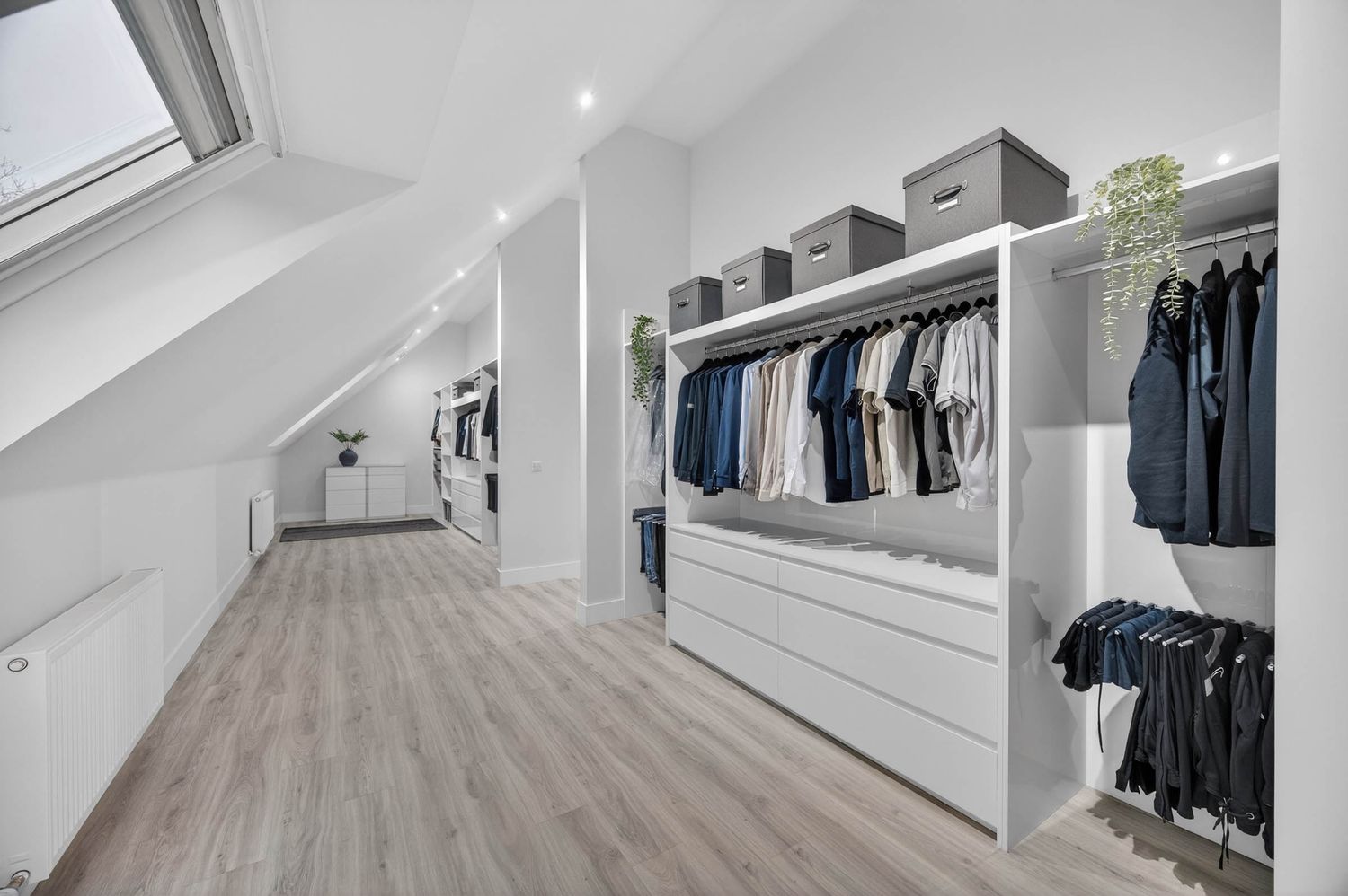 Walk-in closet with white built-in shelving and clothing racks under a sloped ceiling with natural lighting.