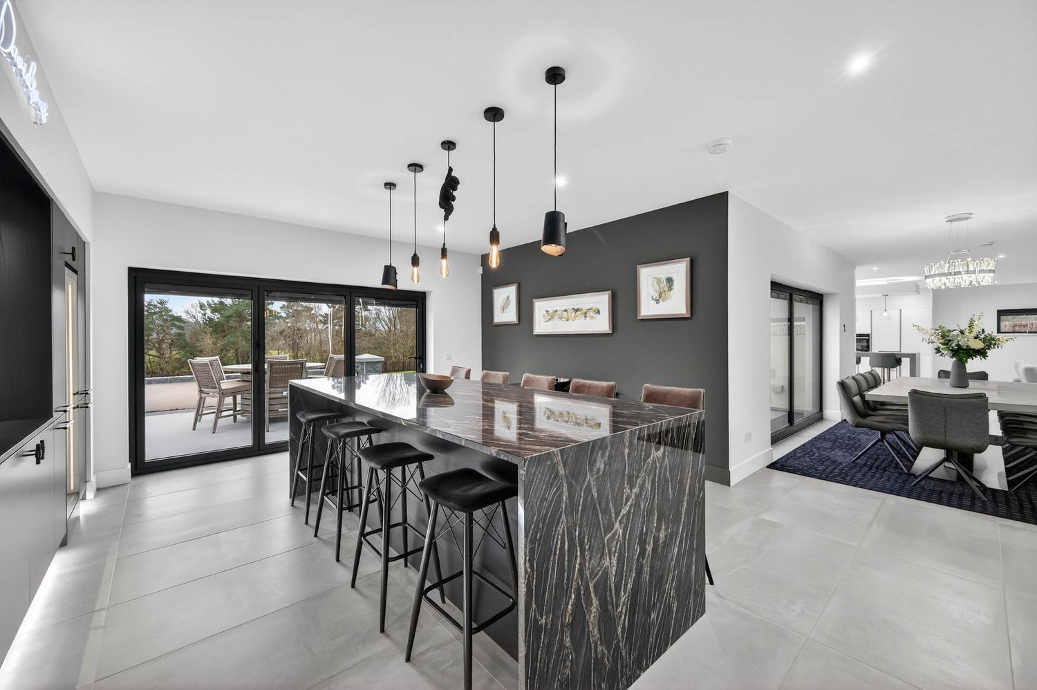Modern open concept kitchen with pendant lights, white cabinets, and black bar stools along a marble island counter.