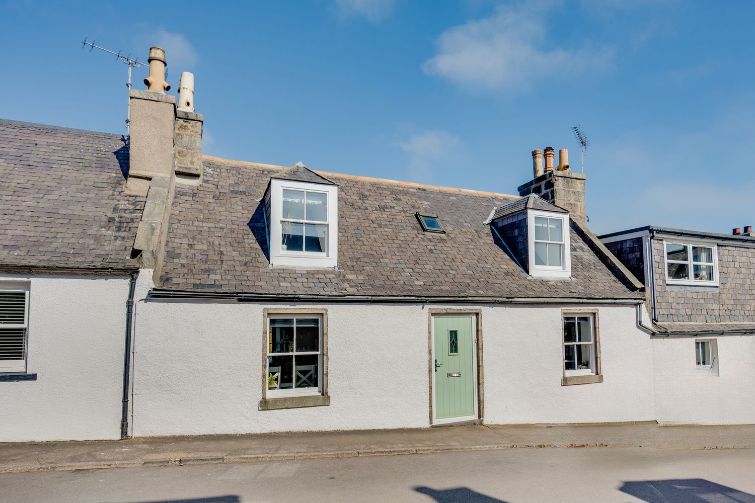 Traditional whitewashed Scottish cottage with mint green door and dormer windows on a sunny day in coastal village.