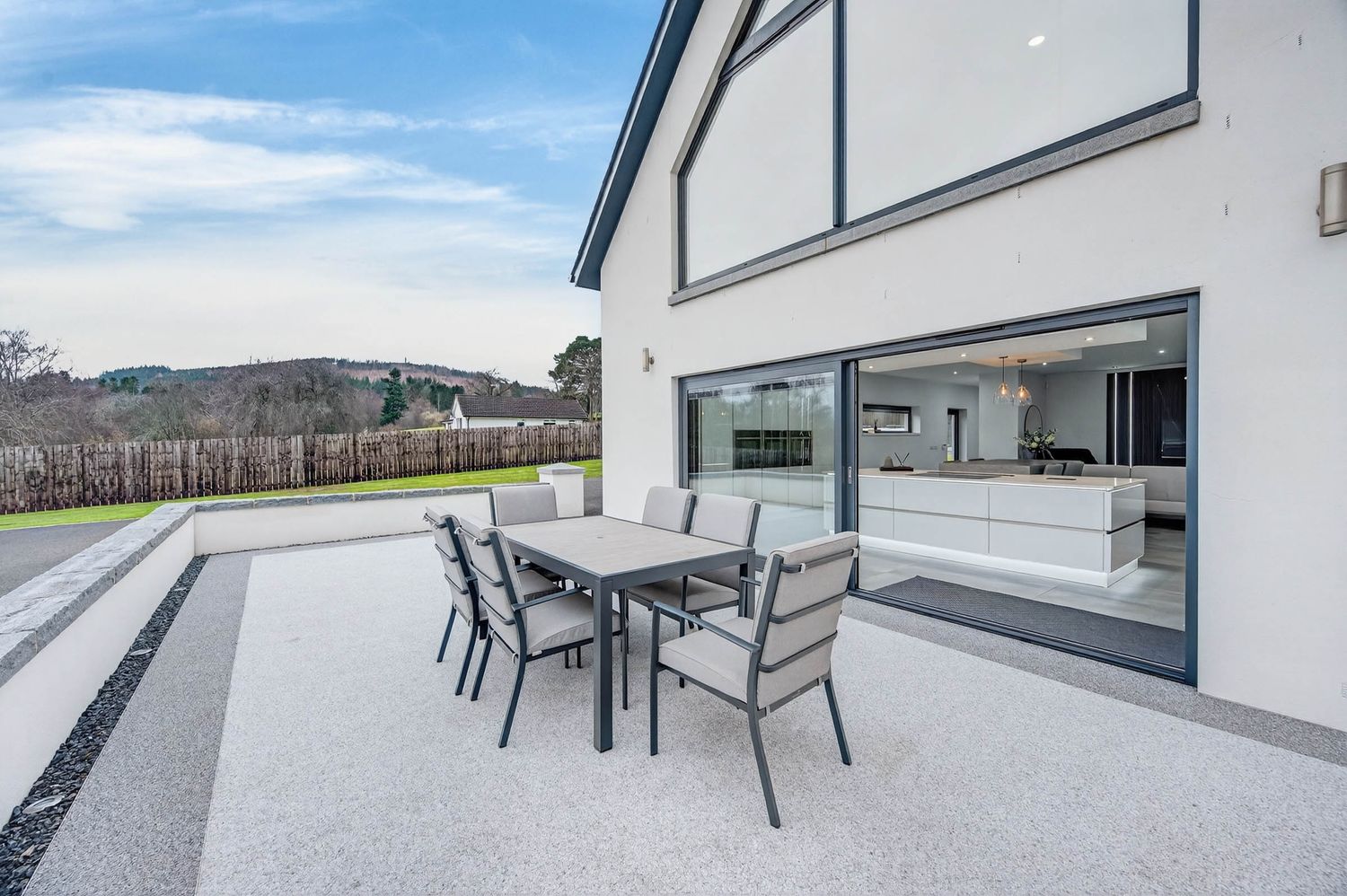 Modern white house exterior with glass sliding doors and outdoor dining set on concrete patio overlooking scenic landscape.