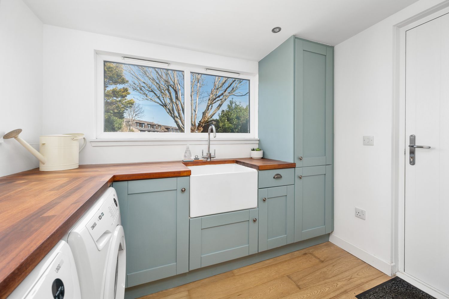 Modern utility room with sage green cabinets, wooden countertops, farmhouse sink and large window overlooking trees.