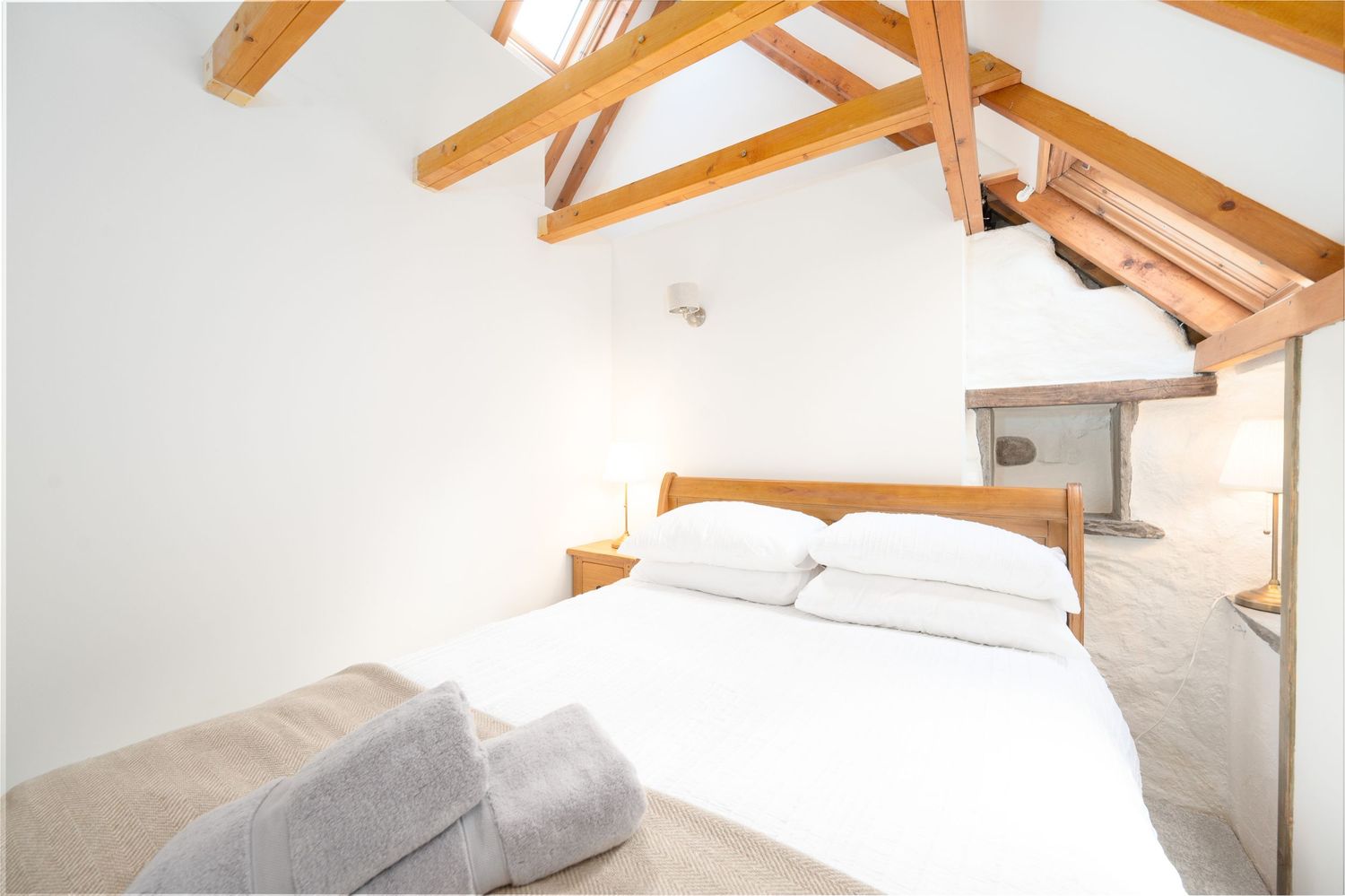 A white bedroom interior with exposed wooden beams and sloped ceiling features crisp linens and minimalist decor.