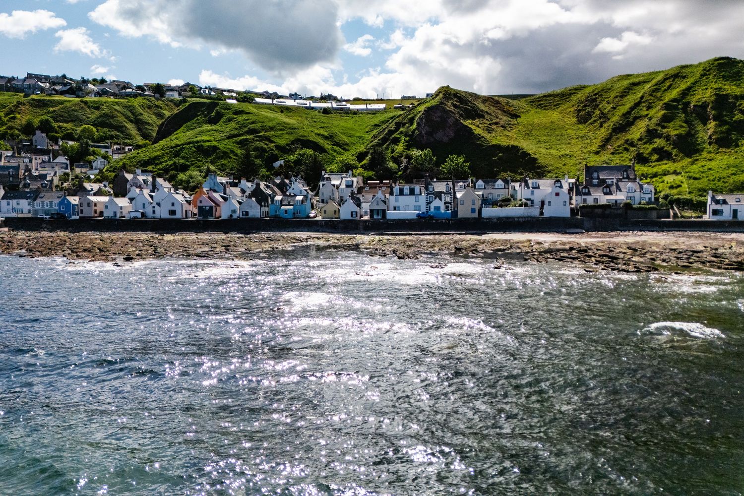 Seaside village with white houses clustered along rocky coastline beneath green rolling hills.