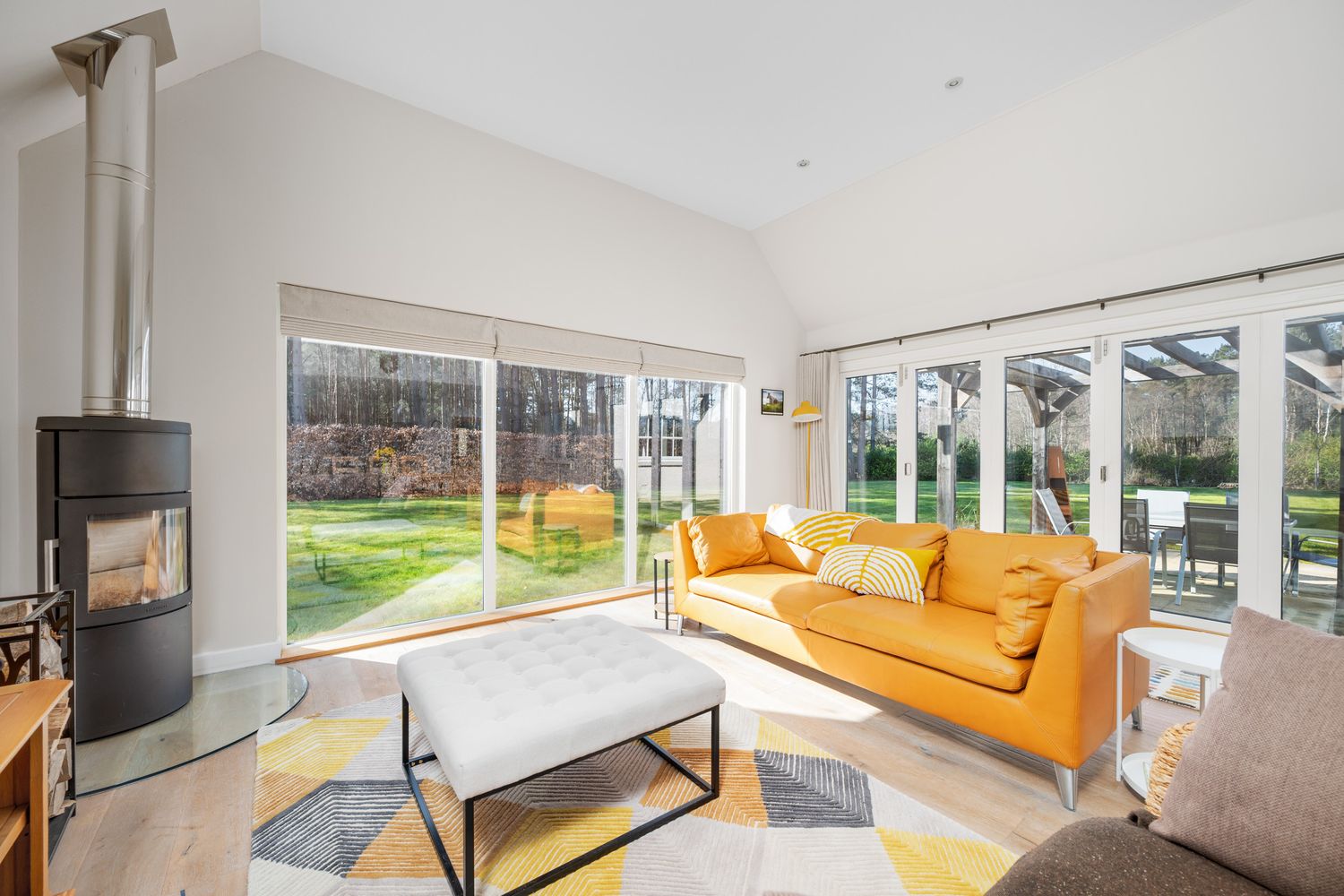 Modern living room with yellow sofa, wood stove, and large windows overlooking garden landscape.