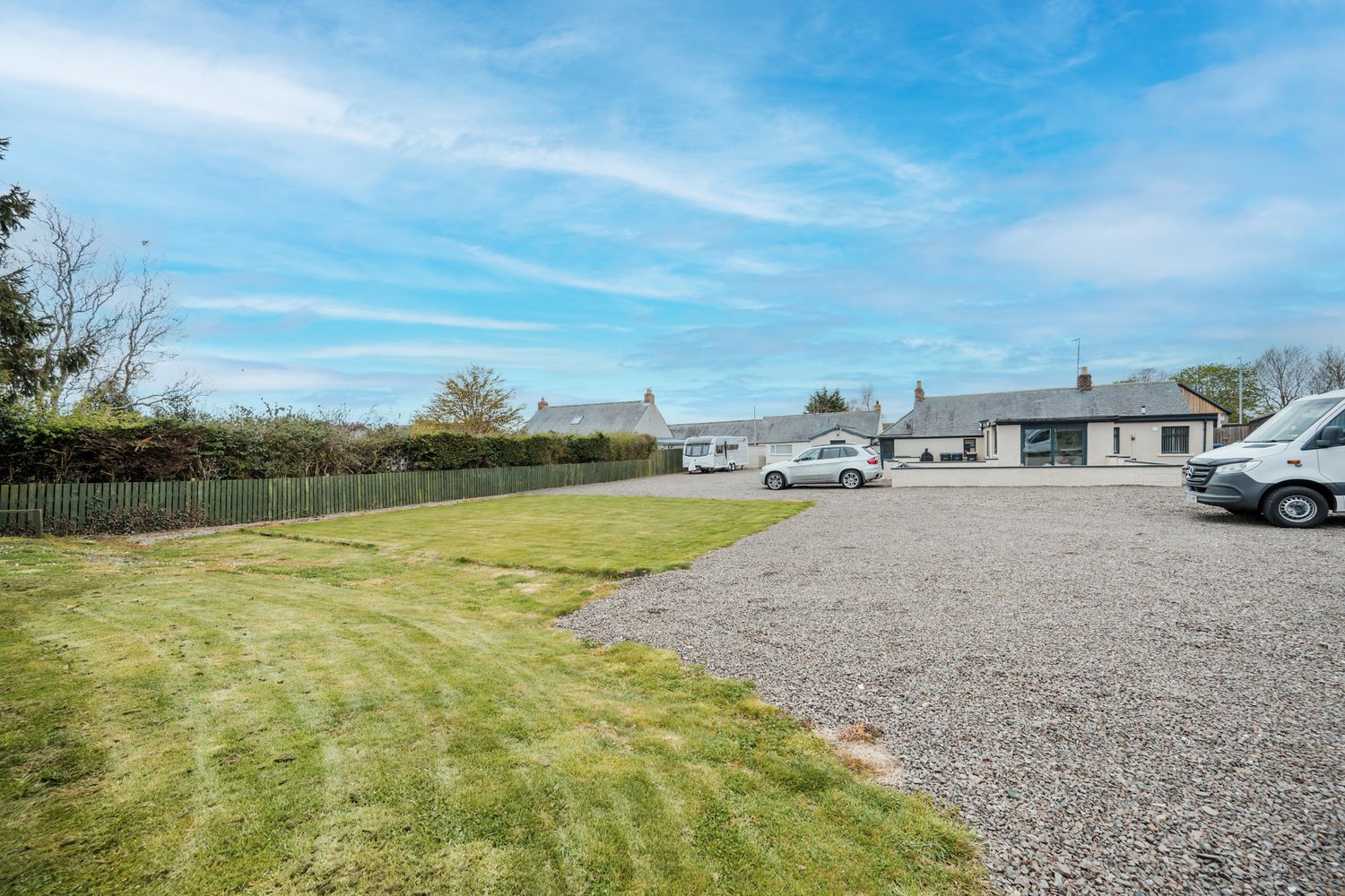Large gravel parking area next to a manicured grass lawn with residential buildings in the background under blue skies.