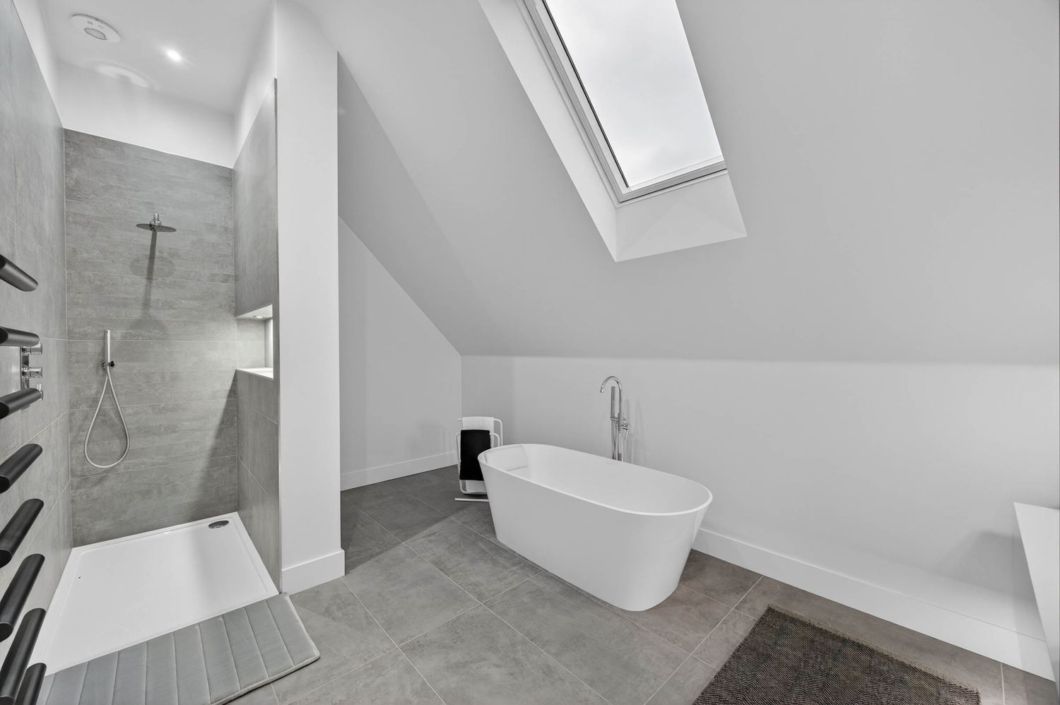 Modern white bathroom with freestanding tub, walk-in shower, and skylight in sloped ceiling features gray tile flooring.