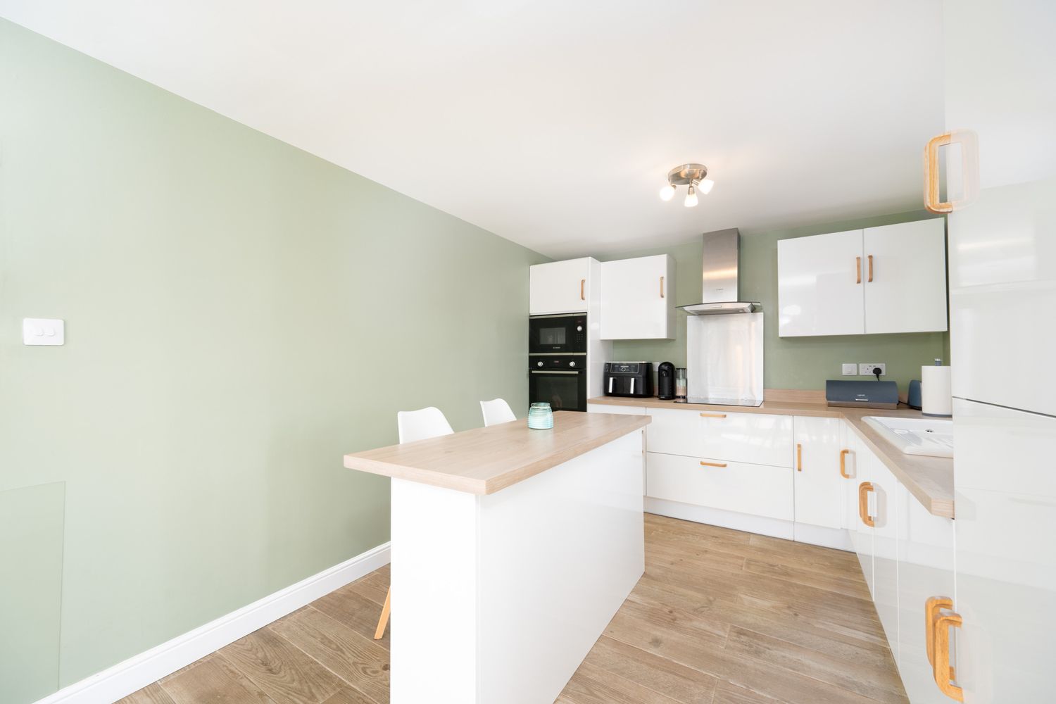 Modern L-shaped kitchen with white cabinets, wood accents, sage green wall and stainless steel appliances.