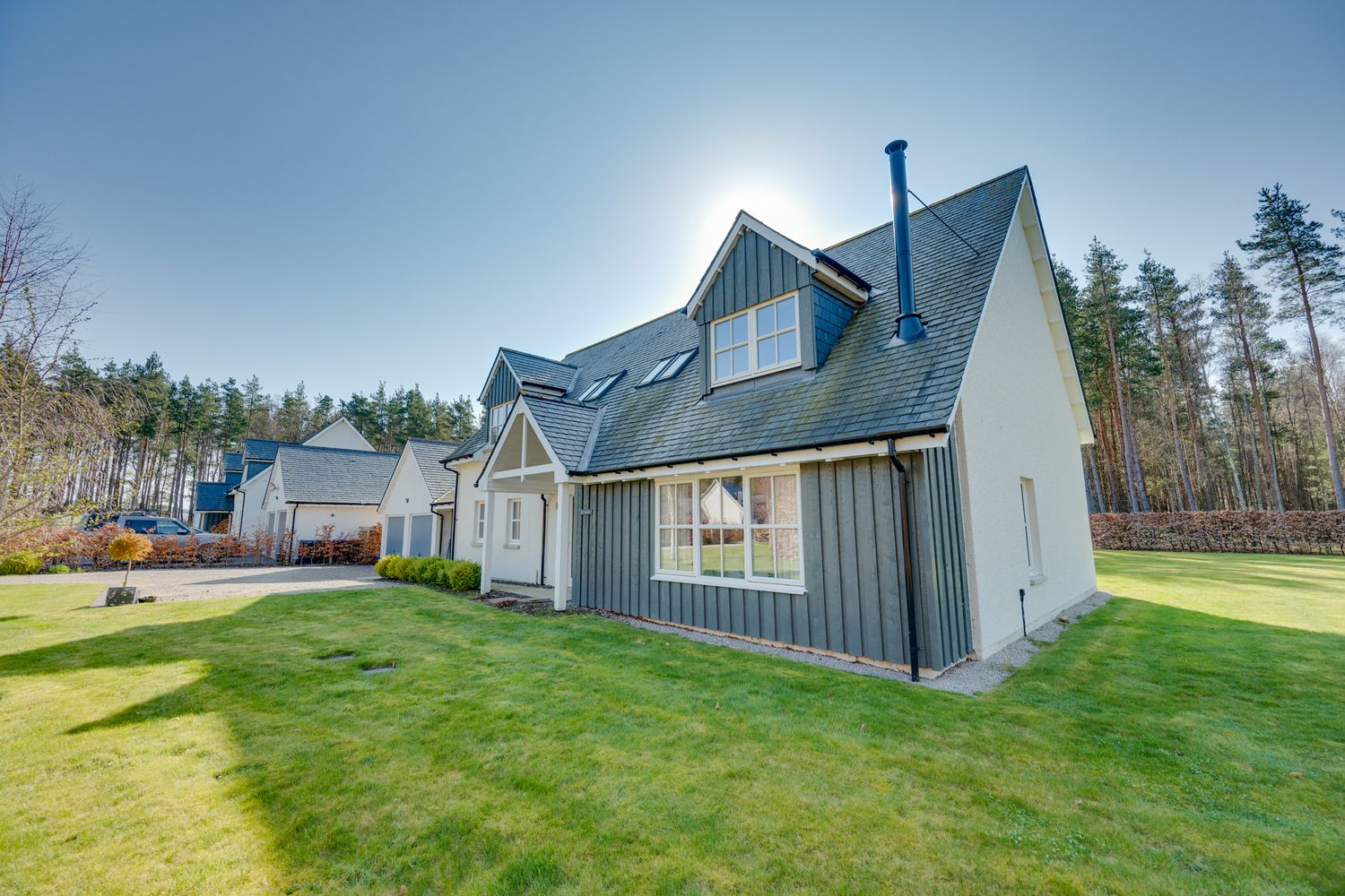 Modern gray and white country house with steep rooflines set against evergreen trees and blue sky.