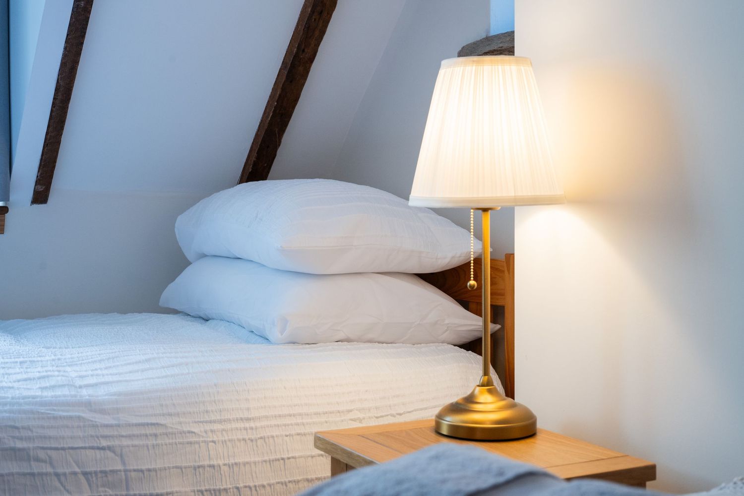 Bedside table with brass lamp next to white pillows under sloped ceiling with exposed beams.