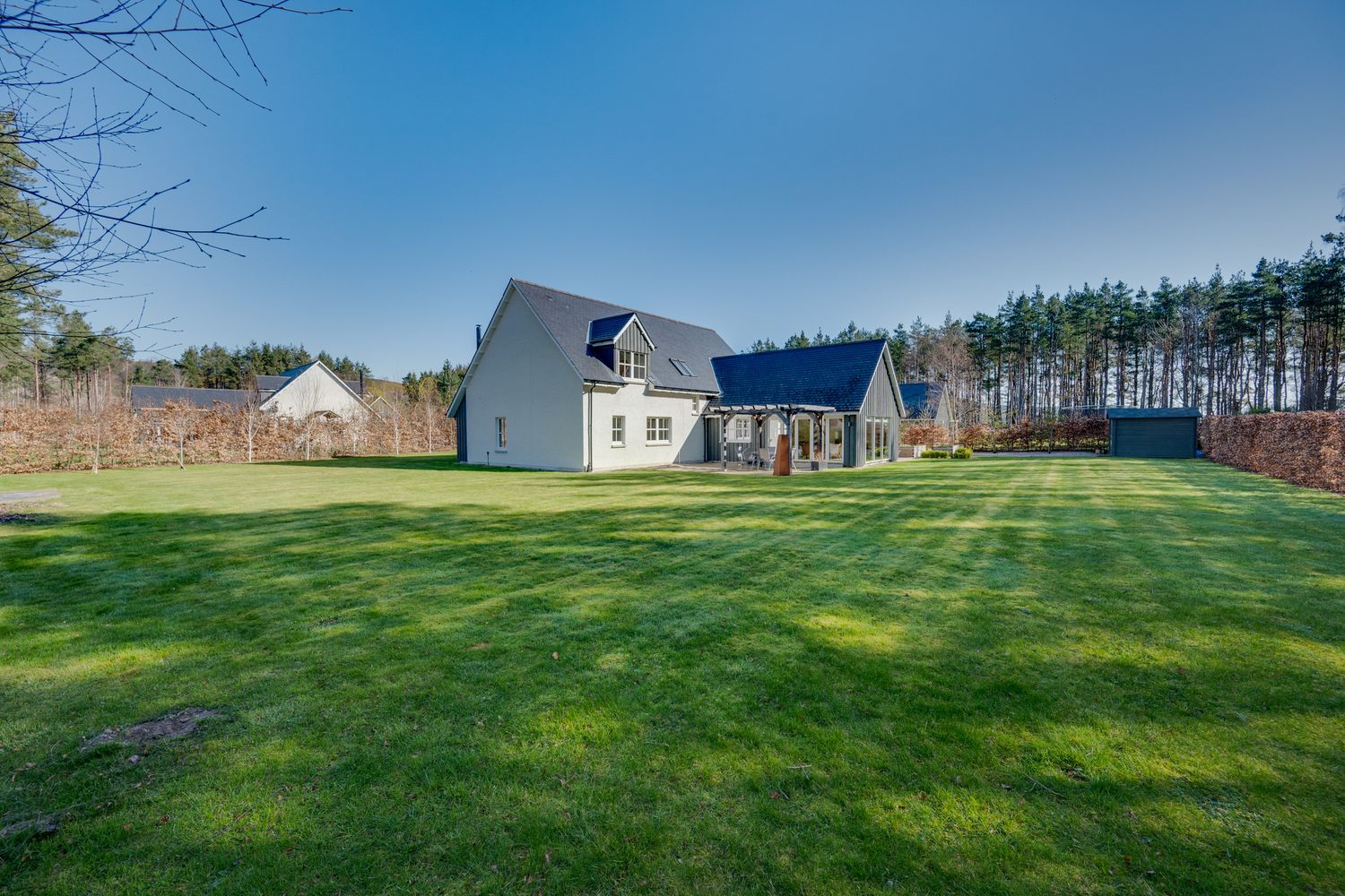 Large white house with attached garage sits on expansive green lawn bordered by pine trees on a sunny day.