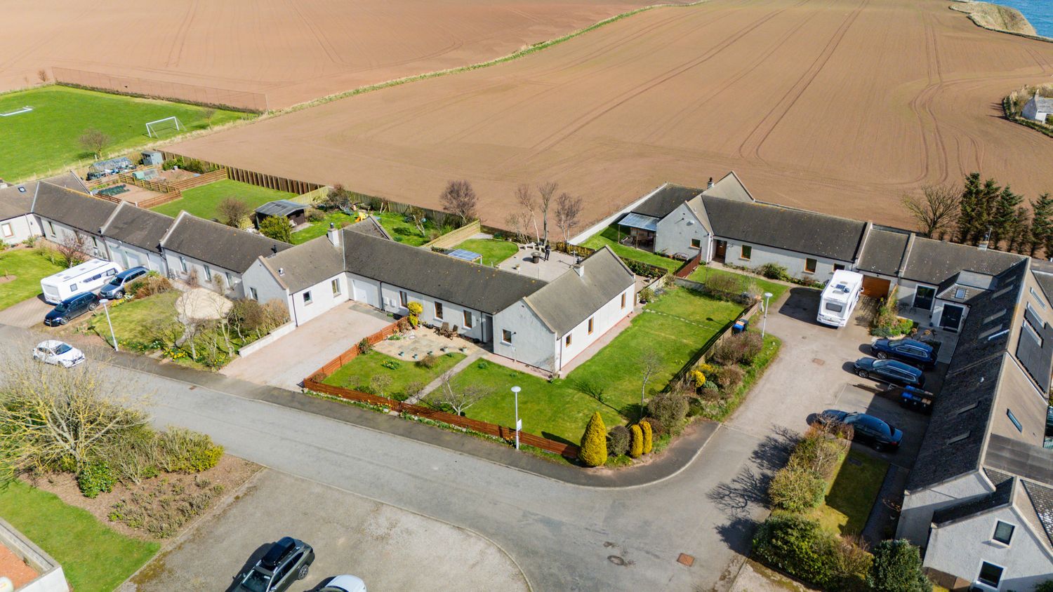 Drone view of white residential buildings with neat gardens arranged around a corner intersection in a rural setting.