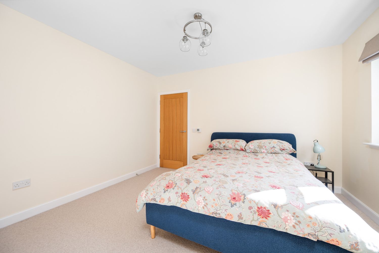 A bright bedroom with a blue bed frame, floral bedding, and wooden door against cream colored walls.