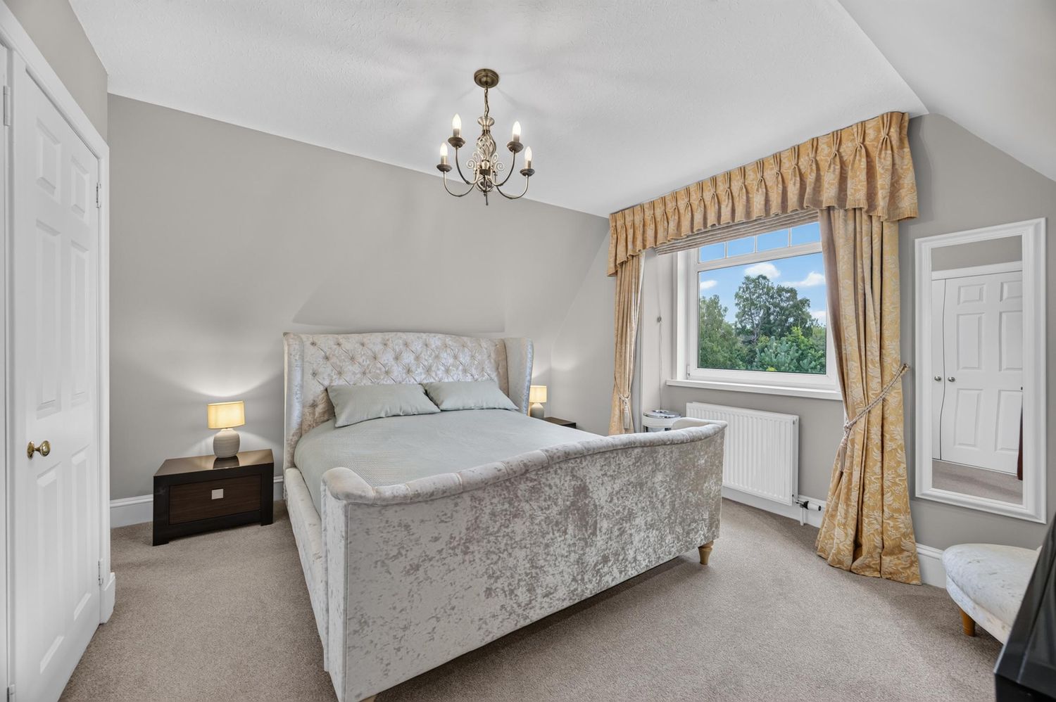 Elegant bedroom with tufted velvet bed, gold curtains, chandelier, and neutral carpet in a bright, well-decorated space.