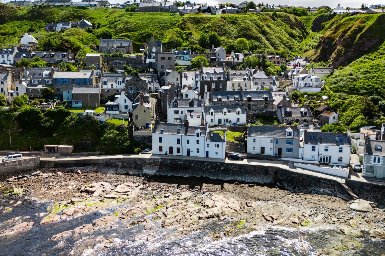 Coastal village with white houses clustered along rocky cliffs and green hillsides in a scenic seaside setting.