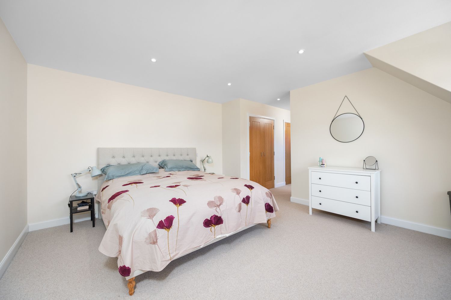 Modern bedroom with grey upholstered headboard, floral bedding, white dresser and circular mirror in neutral tones.