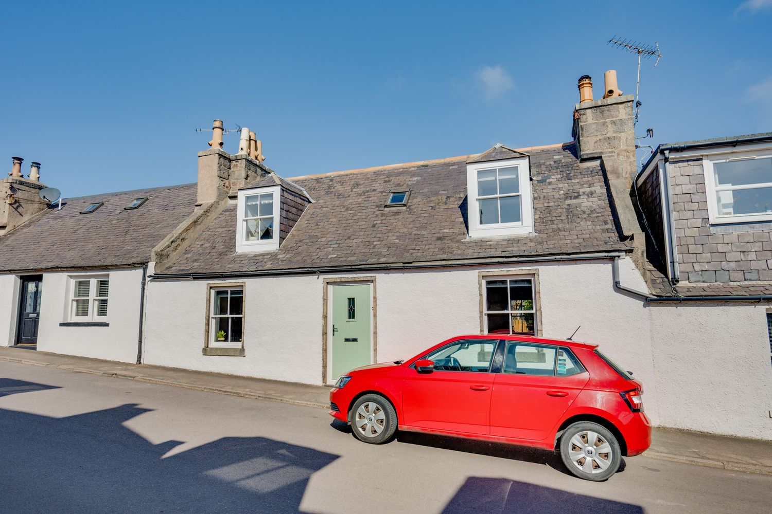 A bright red modern hatchback car parked in front of traditional white Scottish cottages with dormer windows.
