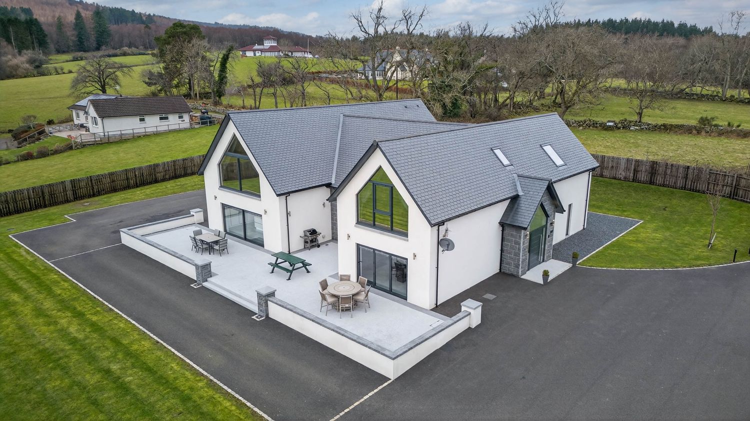 Aerial view of a contemporary white house with grey roof situated on a landscaped rural property with paved driveway.