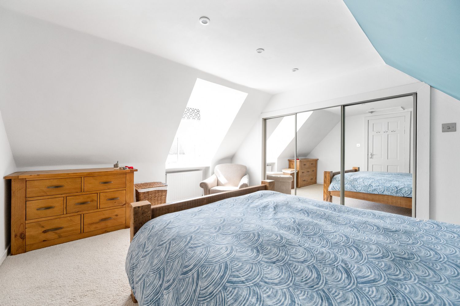 Modern loft bedroom with blue bedding, wooden dresser and mirrored wardrobe under sloped ceiling.