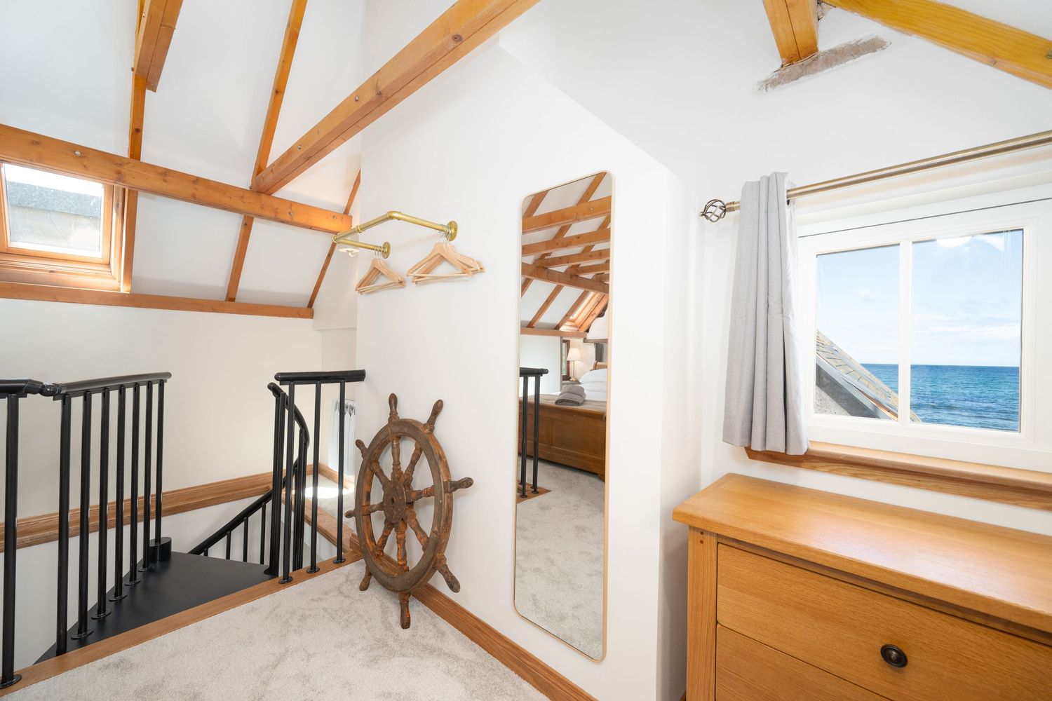 Modern coastal home interior with exposed wooden beams, white walls, and nautical ship wheel decor on the wall.