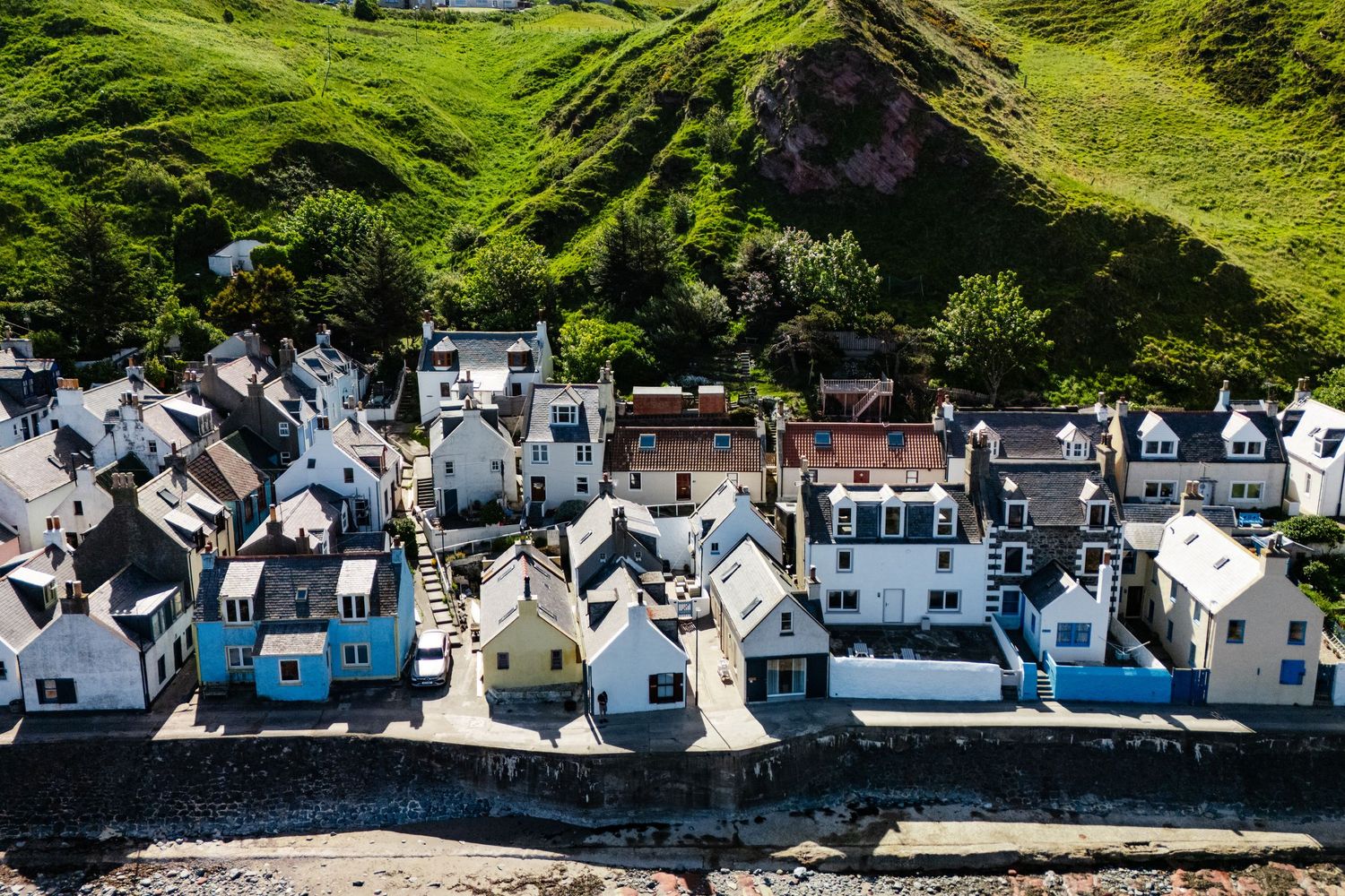 Aerial view of white seaside cottages lined along curved harbor wall with green hills above.