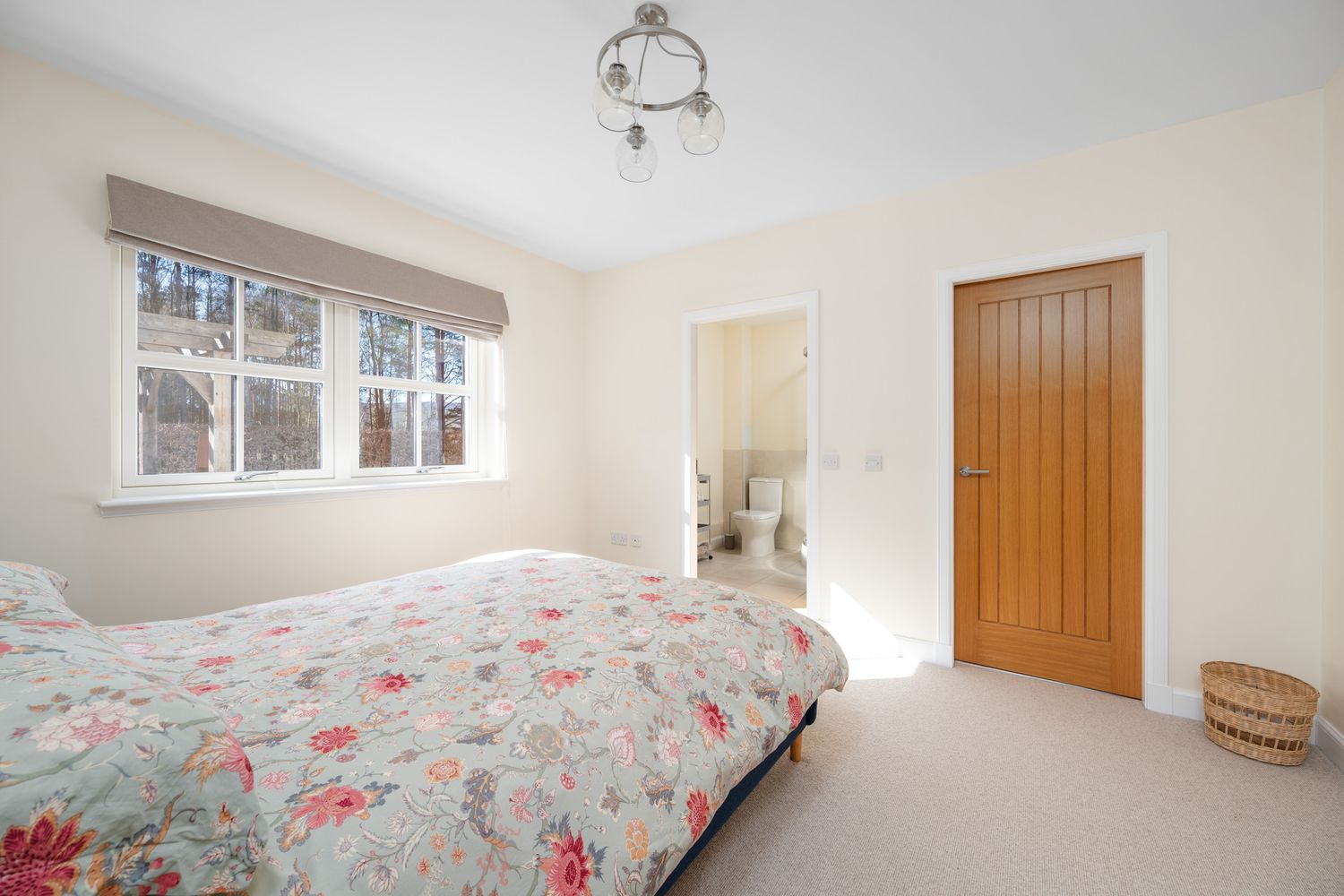Bright modern bedroom with floral bedding, beige walls, window, ensuite bathroom and wooden door.