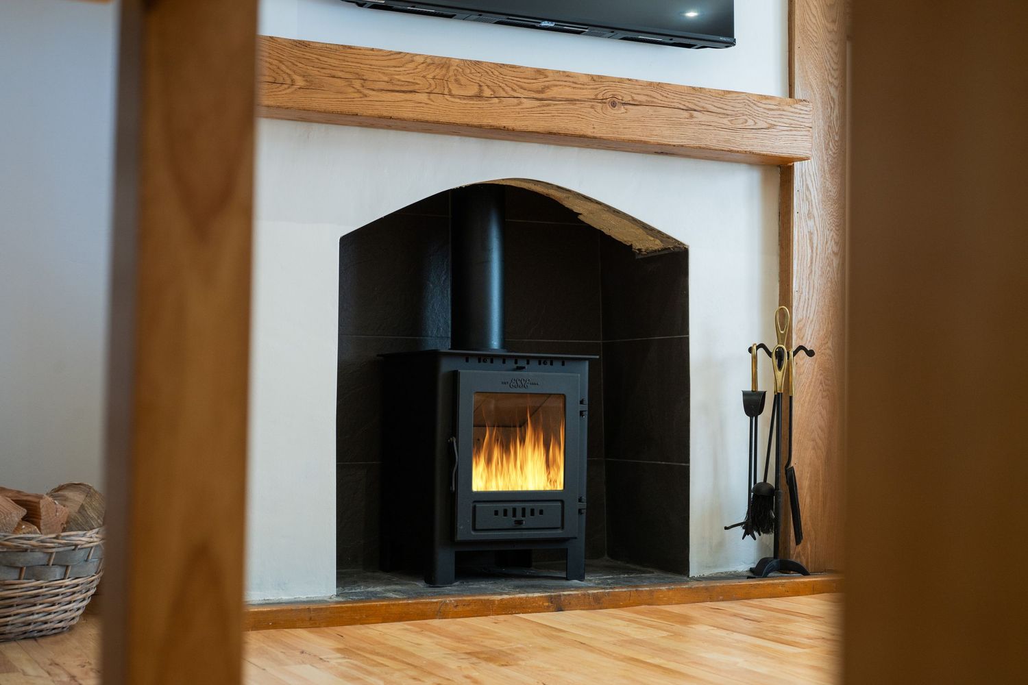 A modern black wood burning stove with glowing flames inside a wooden fireplace surround and fireplace tools nearby.
