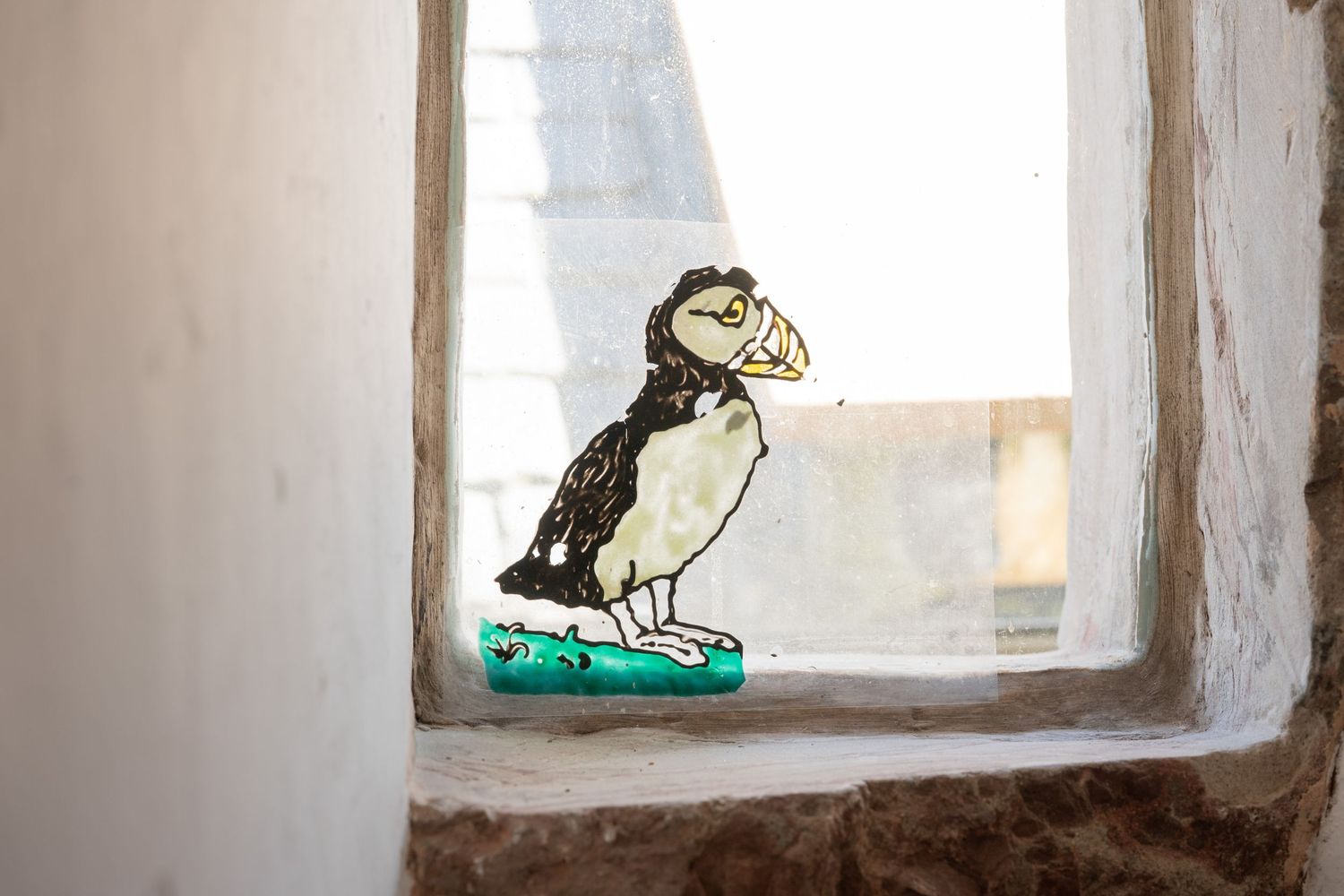 Hand-painted whimsical puffin artwork perches on a windowsill against sunlit white stone walls.