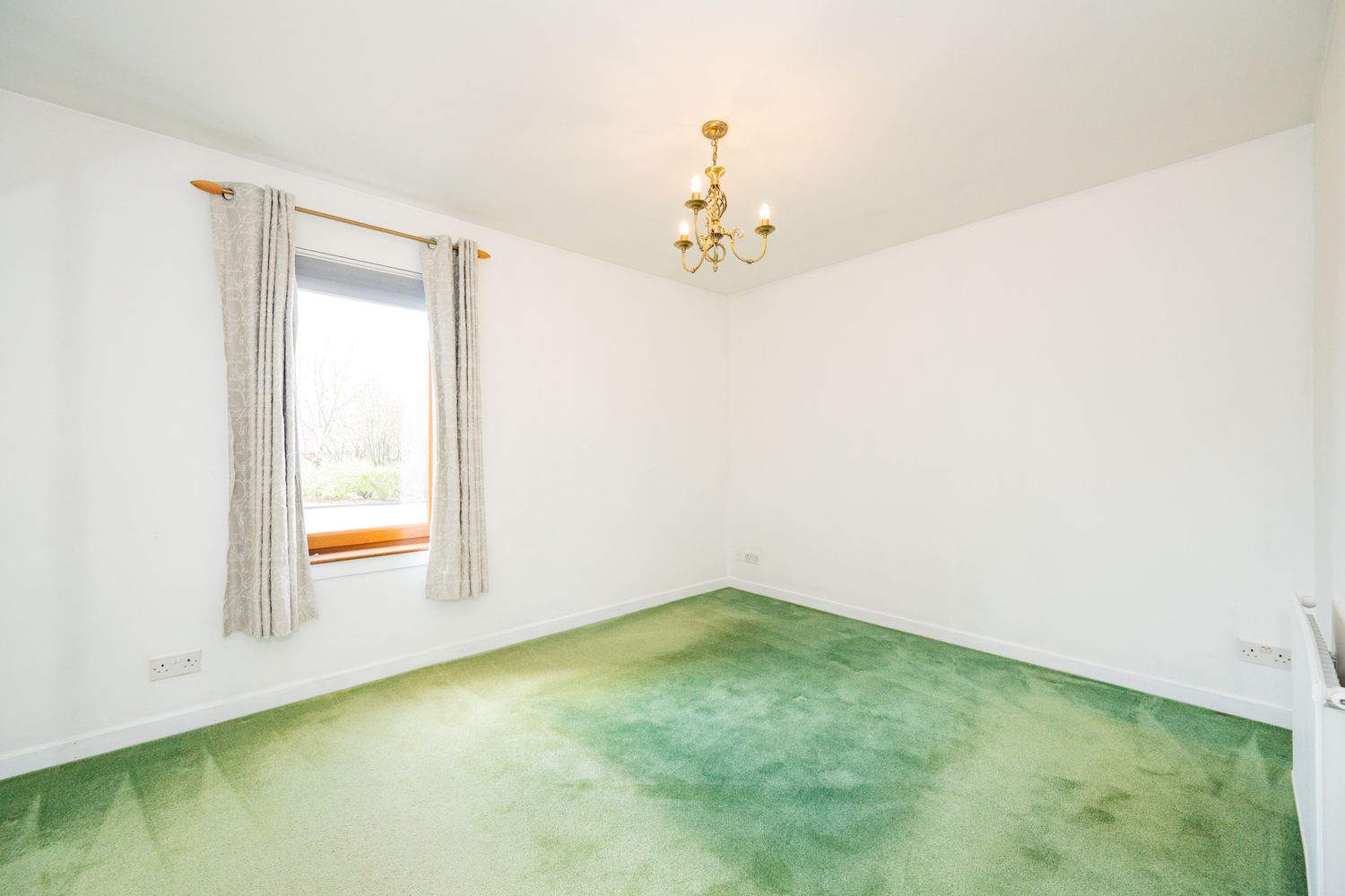 Empty room with green carpeted floor, white walls, single window with curtains, and a basic ceiling light fixture.