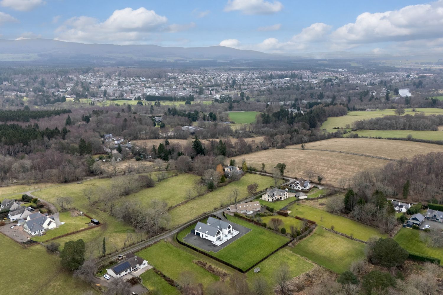 Panoramic aerial view of a suburban area with houses dotting green hillsides under a cloudy blue sky.