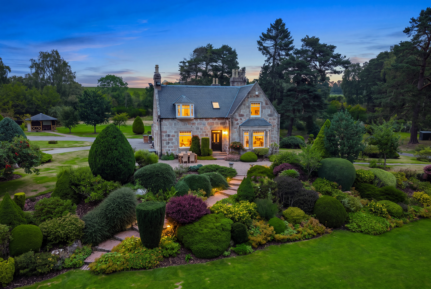 Aerial view of a charming stone cottage with glowing windows surrounded by lush, manicured gardens at dusk.