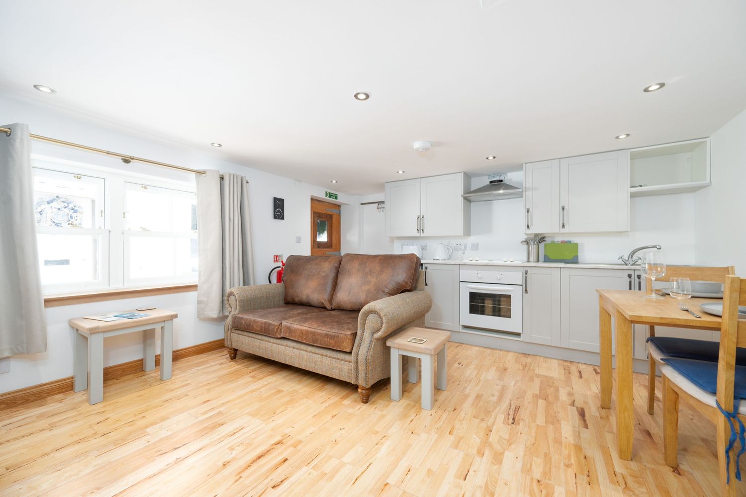 Modern studio apartment interior with open plan kitchen, brown leather sofa and wooden flooring in bright sunlit space.