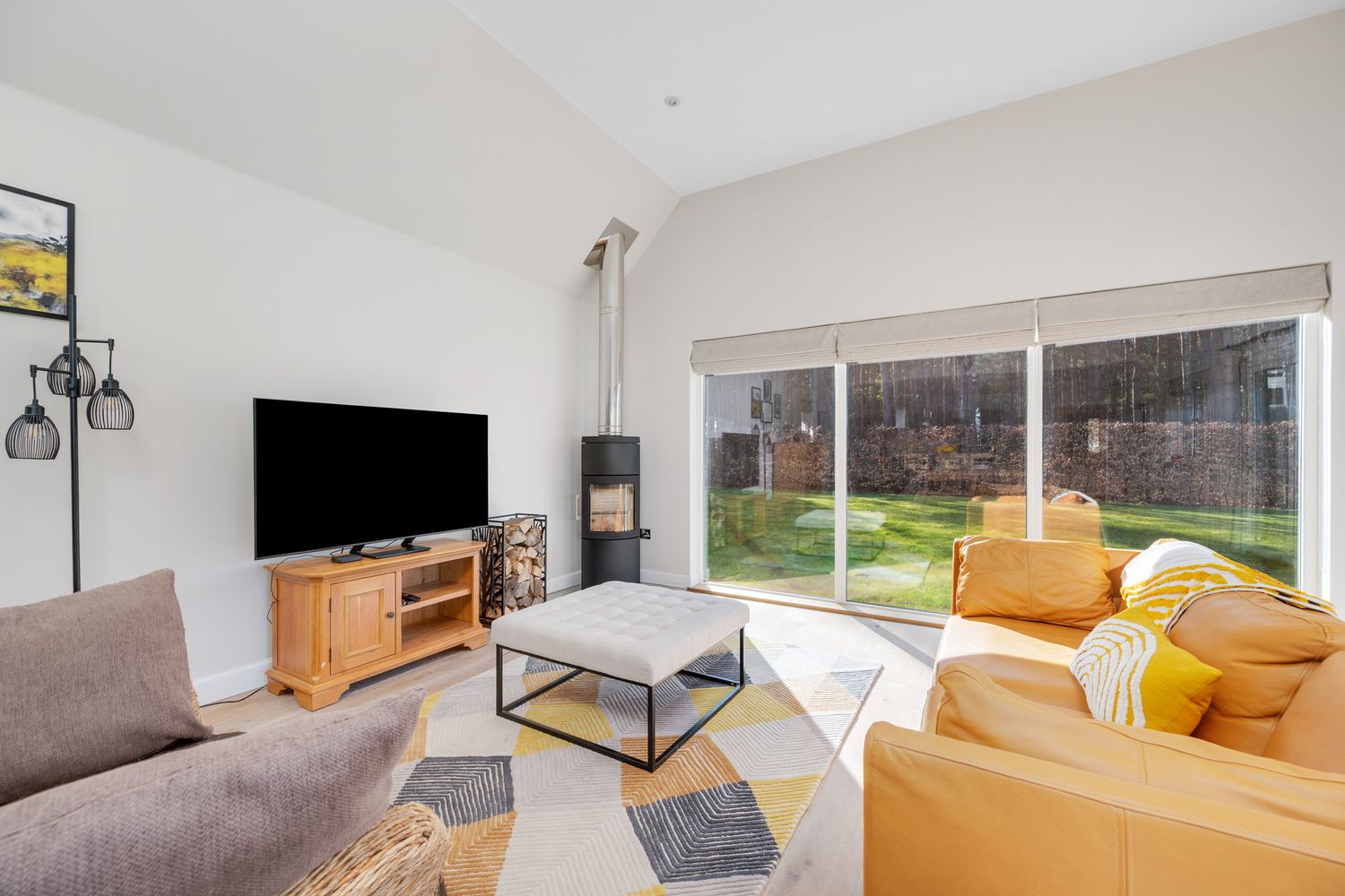 Modern living room with grey and mustard furniture, wood TV stand, and large windows overlooking a garden.